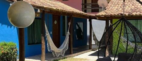 Una terraza de madera con una hamaca y una silla colgante, un edificio azul al fondo y una estructura de techo de paja.