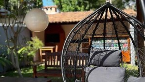 A hanging chair with cushions on a wooden deck, a table, and a lamp post in a garden setting. - Pousada Tropicália (Porto Seguro)