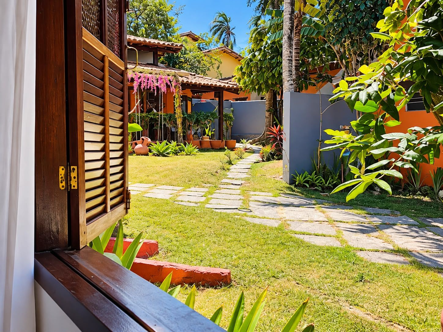 Un sendero de jardín que conduce a una puerta de madera con una ventana, rodeado de exuberante follaje y plantas en macetas.