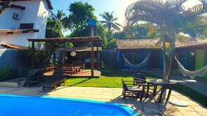 A swimming pool with lounge chairs and a pergola, surrounded by a garden and a small building. - Pousada Tropicália (Porto Seguro)