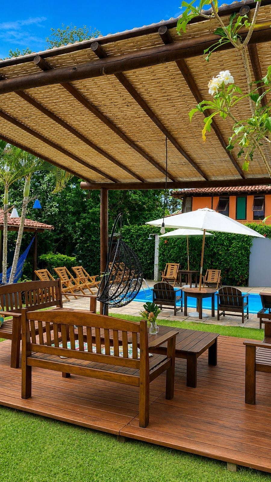 A covered outdoor seating area with wooden furniture, a hanging chair, and a pool in the background.