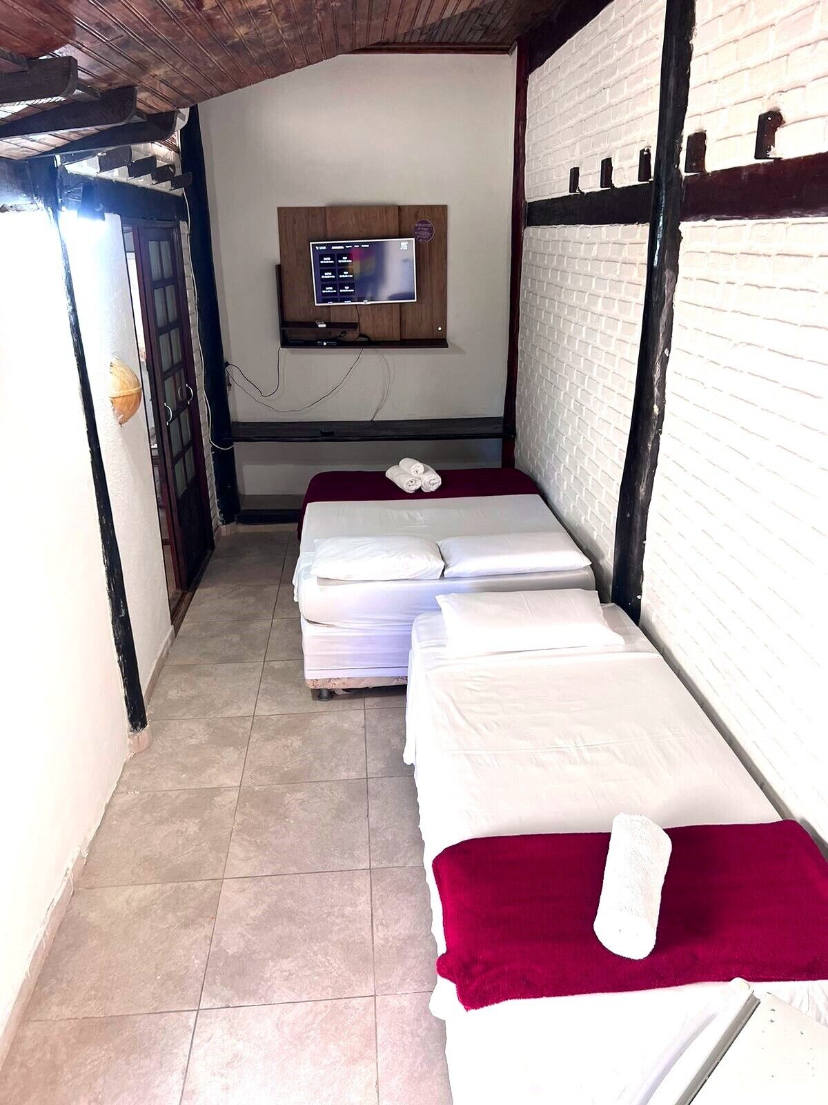 A small, narrow room with two beds, a television mounted on the wall, and a tiled floor.
