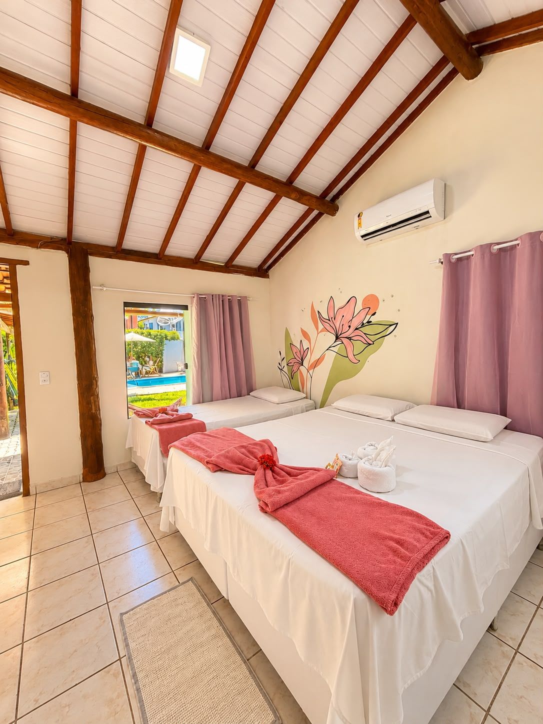 A hotel room with a wooden ceiling, two beds with white linens, a wall-mounted air conditioner, and a view of a pool through a window.