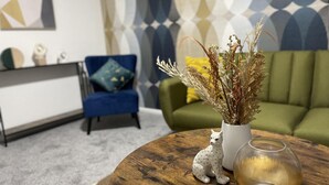 A living room with a round wooden coffee table, a white vase with dried plants, and a leopard figurine. A blue armchair and a green sofa are in the background. - Convenient 3 bd House Salford 7 Guests (Salford)