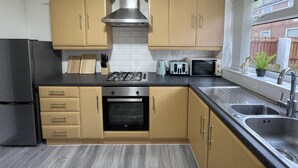 A kitchen with wooden cabinets, a stainless steel range hood, a black refrigerator, a built-in oven, a sink, and a window with a view of a brick wall and a fence. - Convenient 3 bd House Salford 7 Guests (Salford)