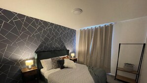 A bedroom with a geometric-patterned wall, a bed with a white quilt, two bedside lamps, a nightstand, a grey throw blanket, a window with curtains, and a black metal shelf unit. - Convenient 3 bd House Salford 7 Guests (Salford)