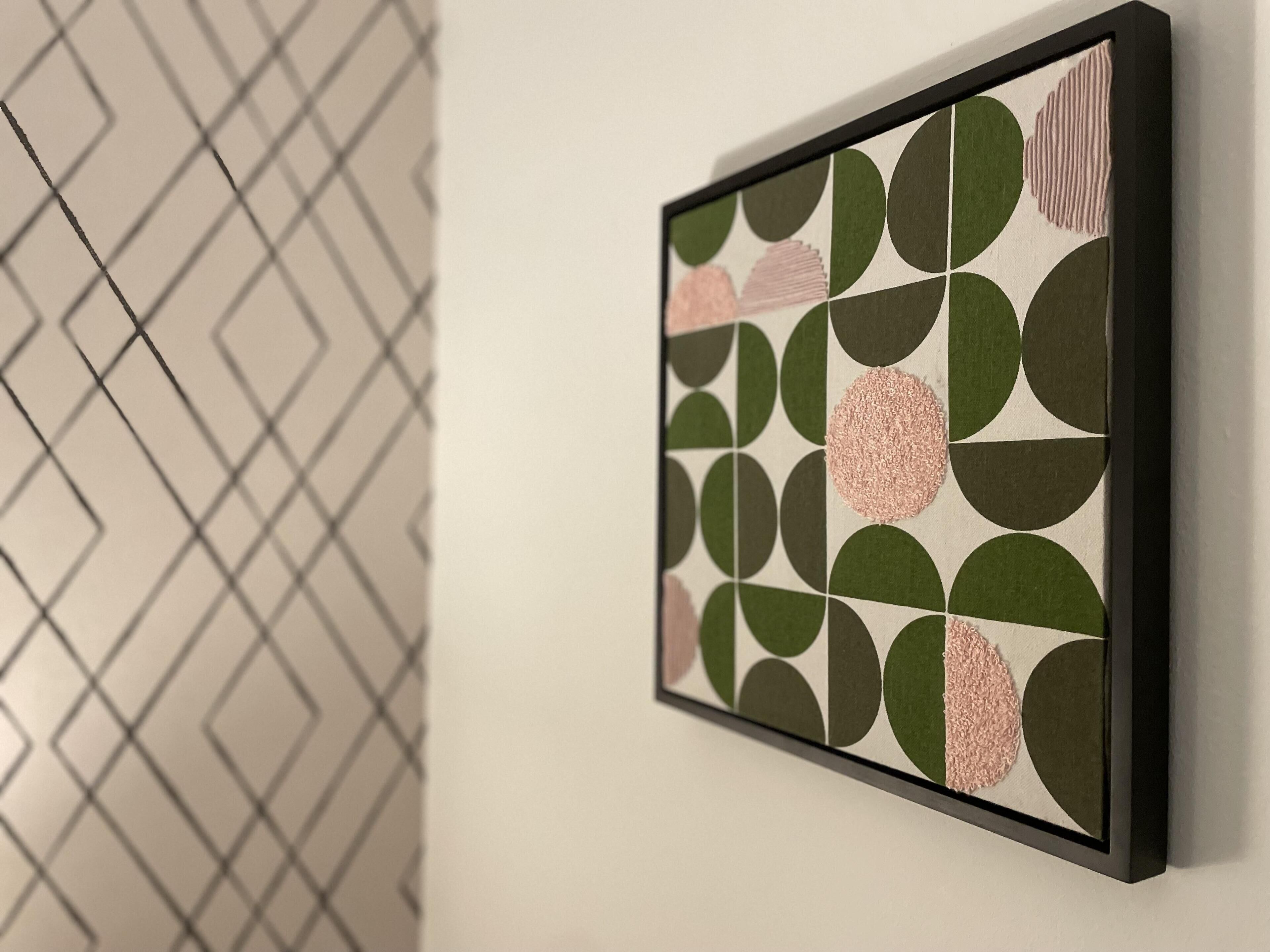 A framed abstract art piece with geometric shapes and a textured circle on a white wall.