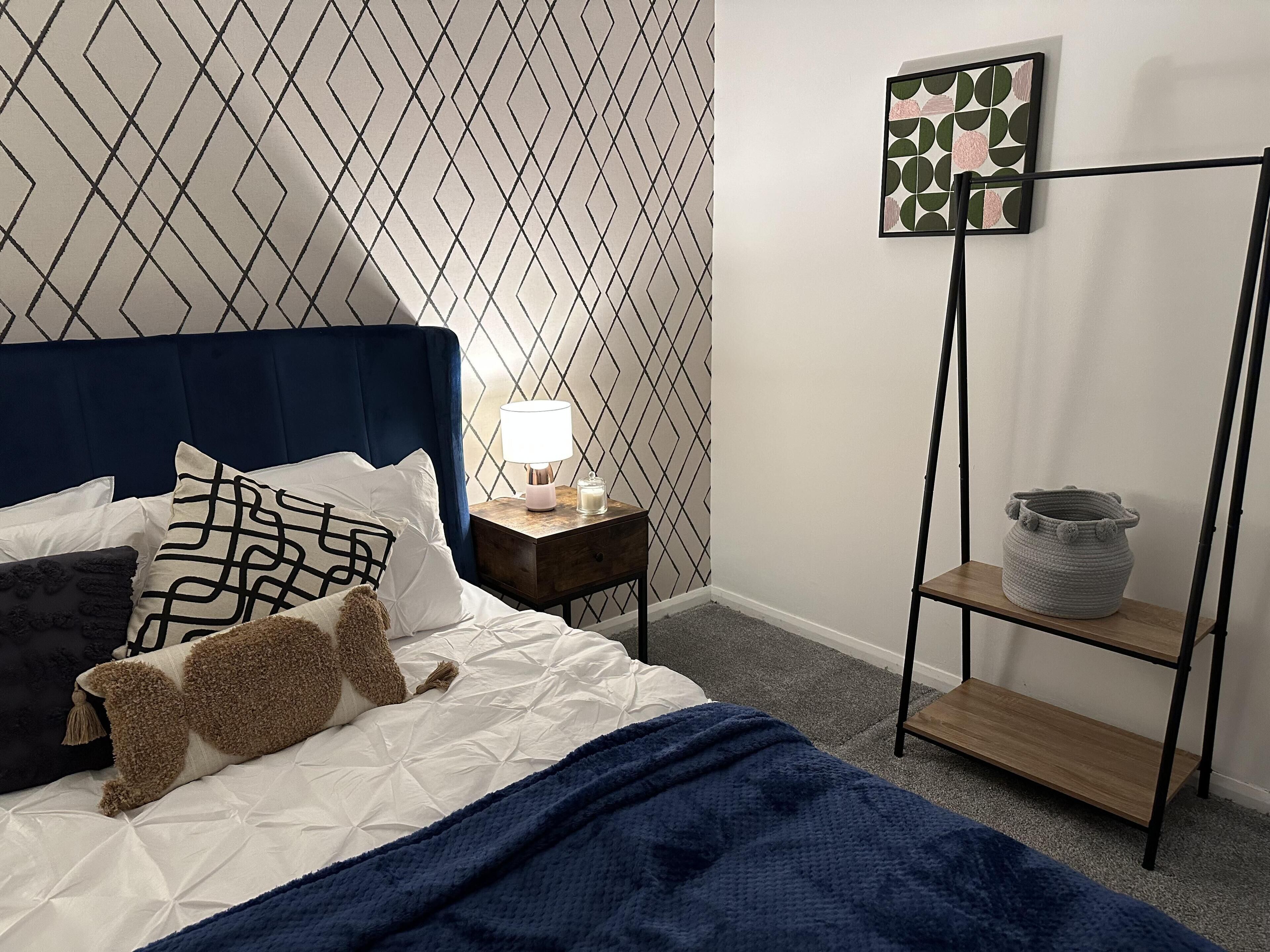 A neatly arranged bedroom with a bed, pillows, a bedside lamp, a wall art piece, and a small shelf with a pot.