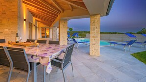 Un espace extérieur aménagé pour les repas, avec un patio couvert, des murs en pierre et une piscine dotée de chaises longues.