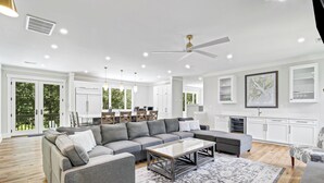 Ein großzügiges Wohnzimmer mit einer grauen Eckcouch, einem Couchtisch und einem gemusterten Teppich. Es gibt eine Essecke mit Tisch und Stühlen sowie eine Küche mit weißen Schränken.