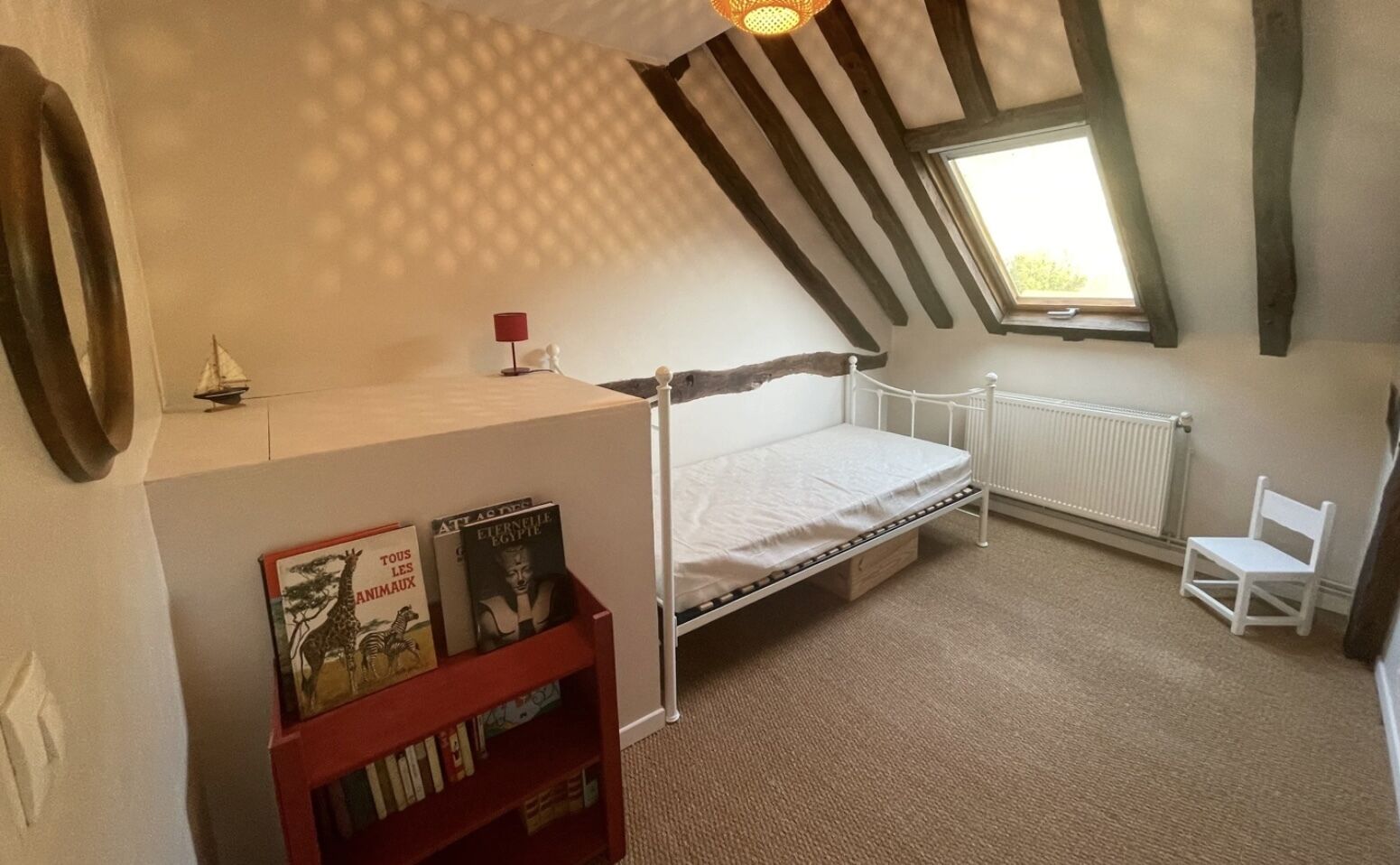 A bedroom with a sloped ceiling, a single bed, a radiator, a small window, and a bookshelf with books.