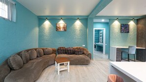 A modern living room with a curved sofa, a small wooden table, and a kitchen area with a white countertop and chairs. - Villa Tiffany (Telavi)