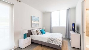 Ein modernes Schlafzimmer mit Bett, Nachttisch, Fernseher und einem großen Fenster.