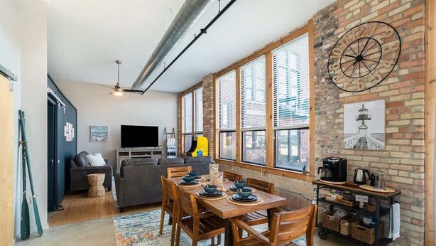 A modern living room with a dining area, a sofa, a TV, and a brick wall with a clock.
