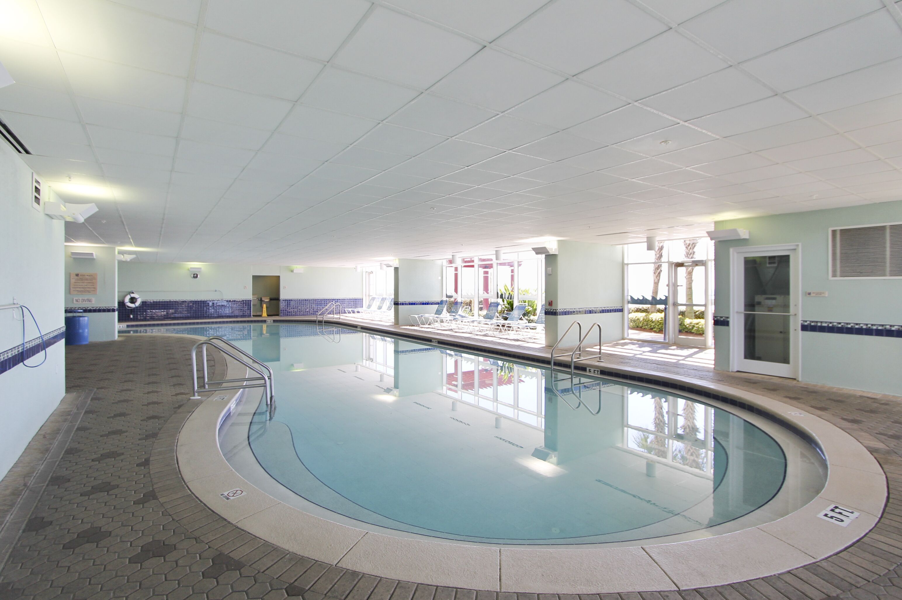 Una amplia piscina interior con suelo de baldosas, una zona de la piscina con sillones de descanso, y una puerta que da al exterior.