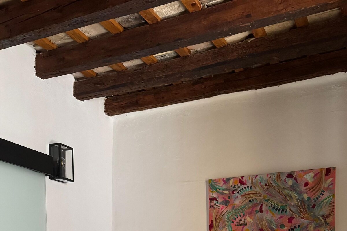 A bedroom with a bed, a painting, a wall-mounted light, and a shelf with a clock.