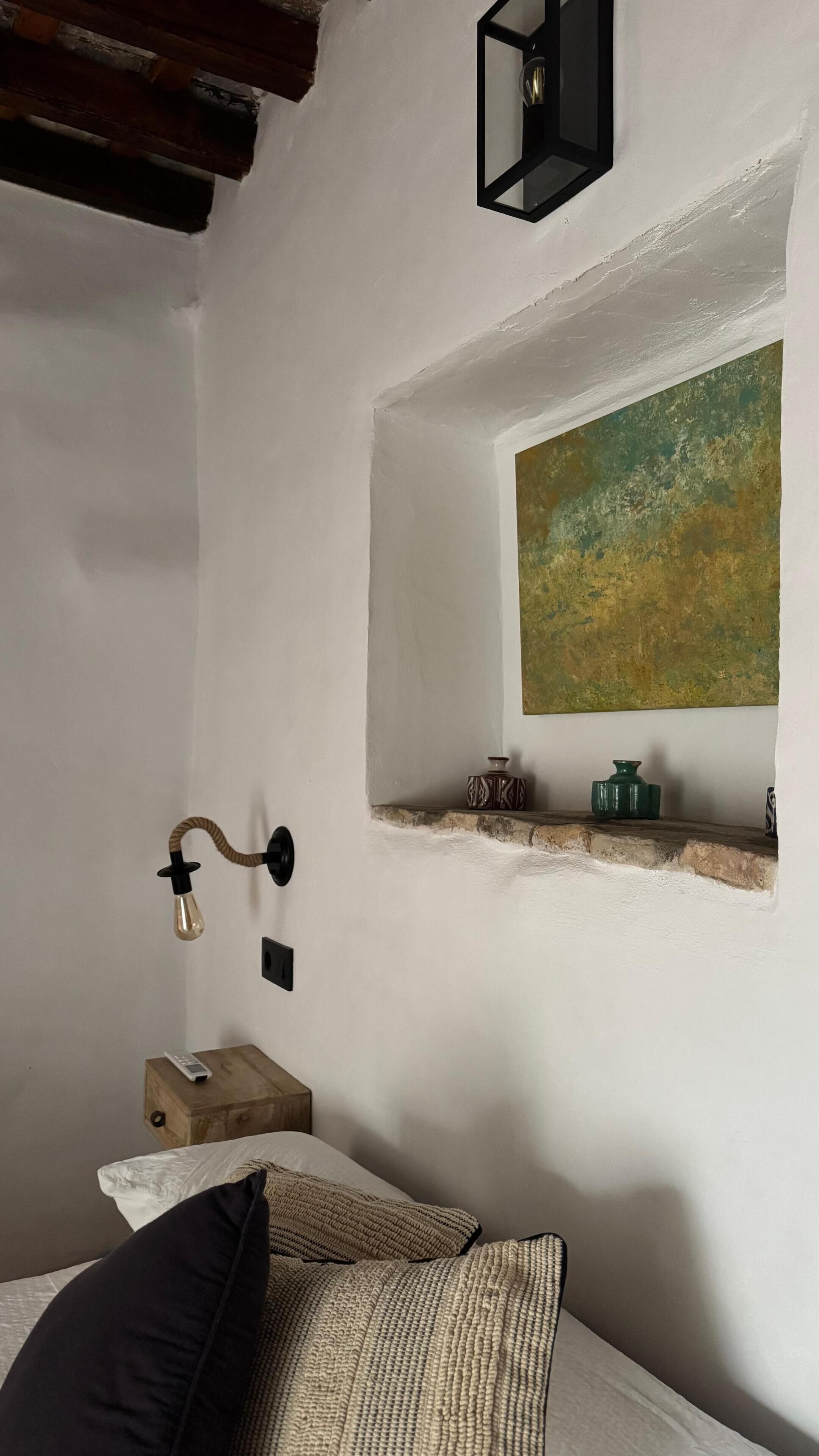 Een slaapkamer met een bed, nachtkastje, lamp, schilderij en een plank met decoratieve objecten.