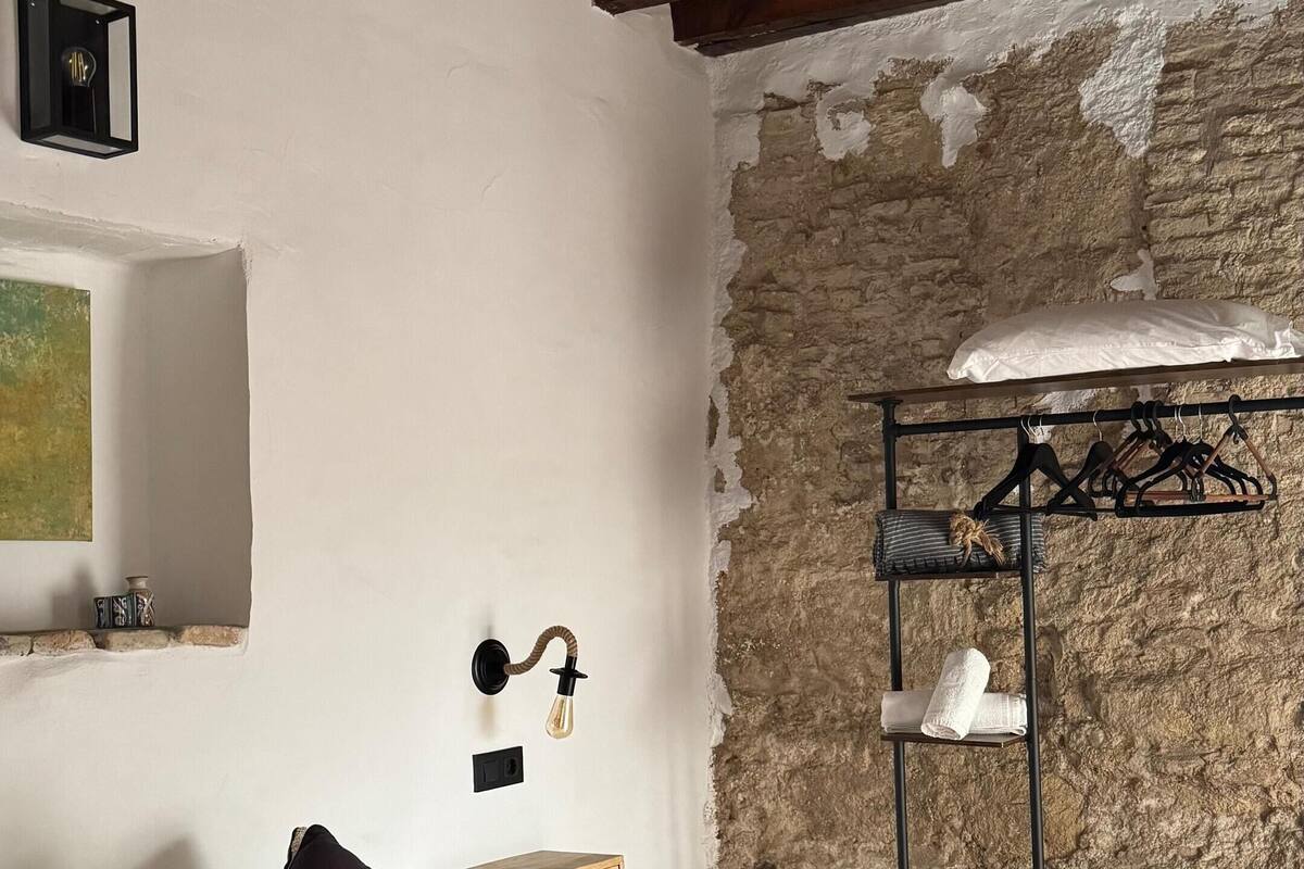 Une chambre à coucher avec un lit, une tête de lit en bois, un mur en pierre, un luminaire suspendu et une étagère avec des vêtements.