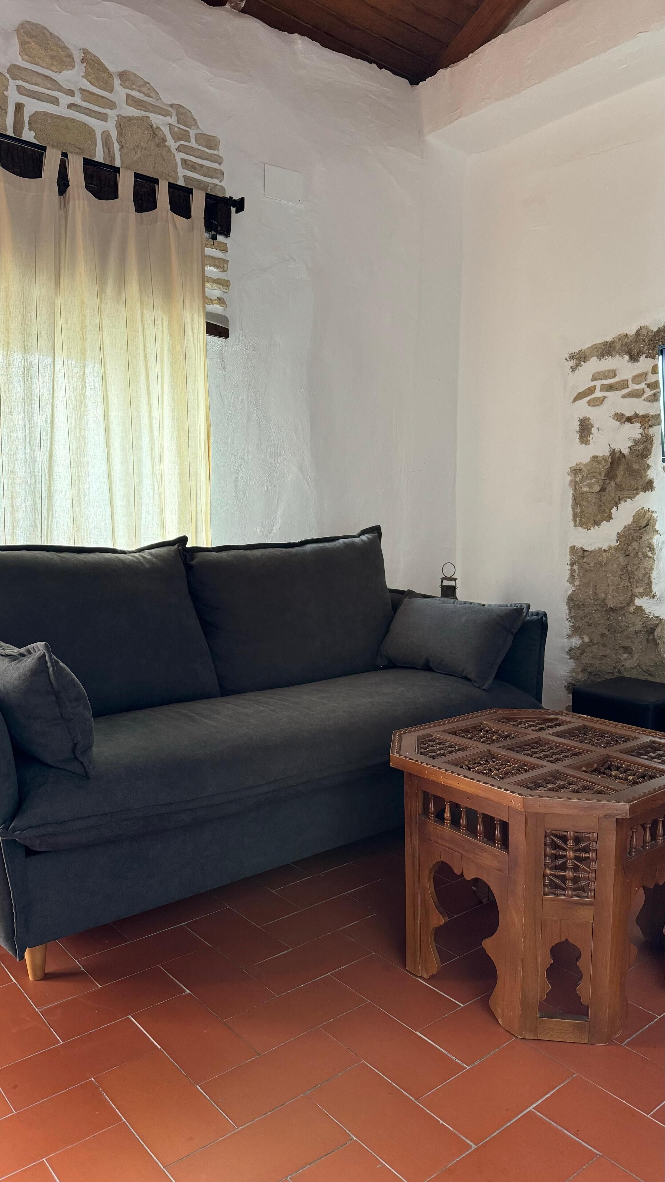 Una habitación con un sofá de color gris oscuro, una mesa de centro de madera con motivos y un suelo de baldosas.