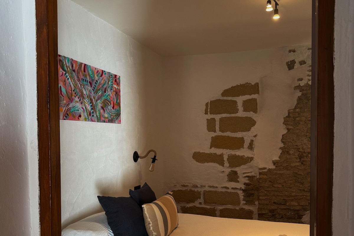 A bedroom with a bed, pillows, a blanket, a wall with exposed brick, and a colorful painting.