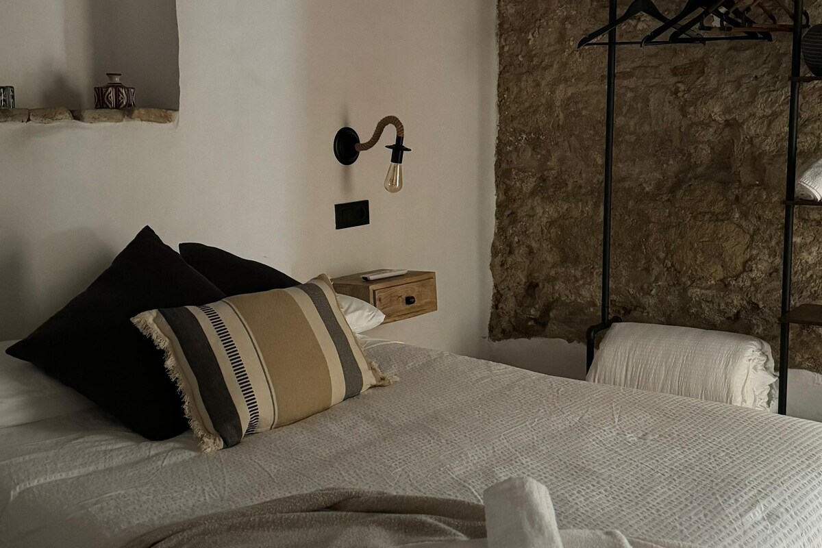 A neatly made bed with a striped pillow, a bedside table with a remote control, and a wall-mounted light fixture.