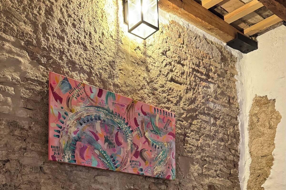 Une pièce au style rustique, avec des murs en briques apparentes, un plafond en bois, un lit recouvert de linge de maison blanc et agrémenté d’oreillers à motifs, une peinture abstraite aux couleurs vives accrochée au mur, et un luminaire suspendu.