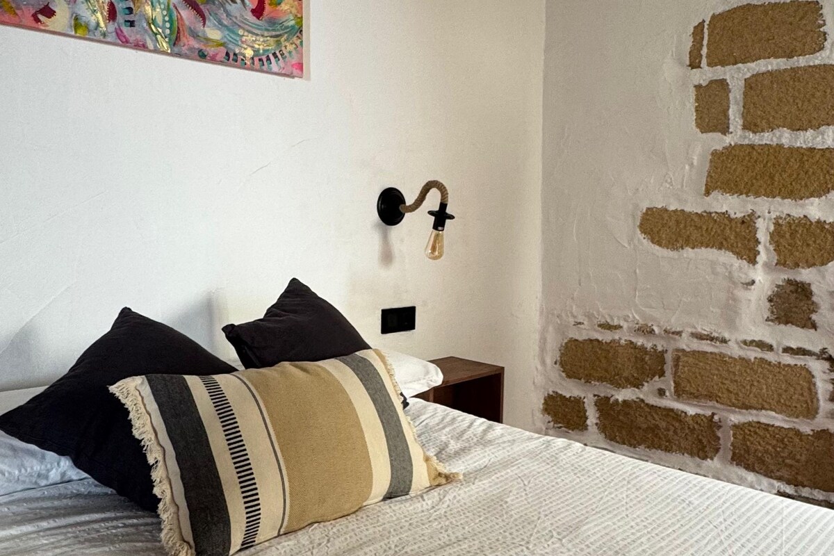 Une chambre comprenant un lit, des oreillers, une œuvre d’art murale, une table de chevet, un luminaire fixé au mur et un mur en briques apparentes.