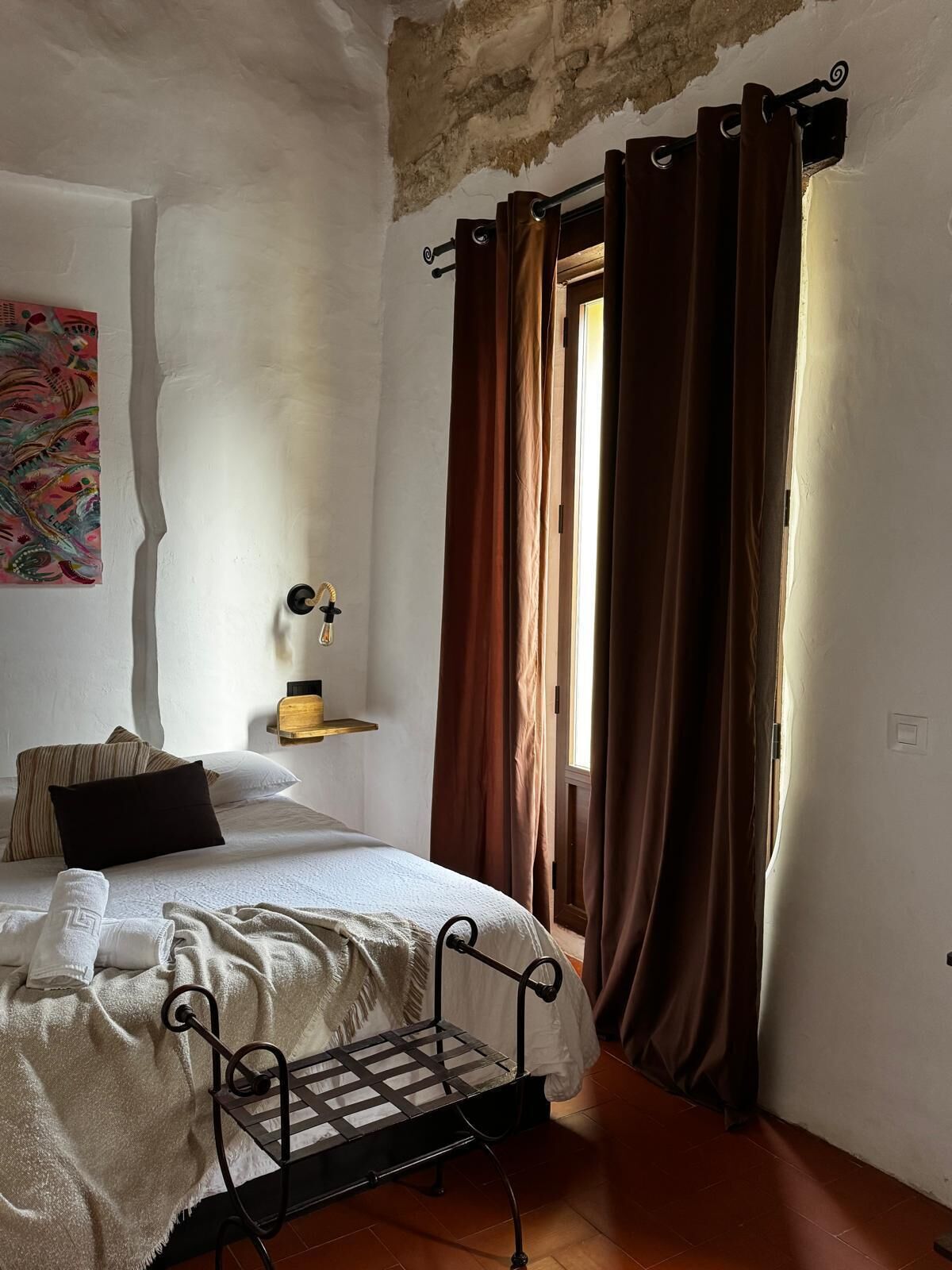 Un dormitorio con cama, mesita de noche, estante en la pared y ventana con cortinas.