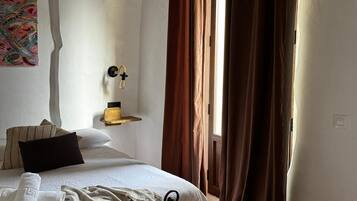 Un dormitorio con cama, mesita de noche, estante en la pared y ventana con cortinas.