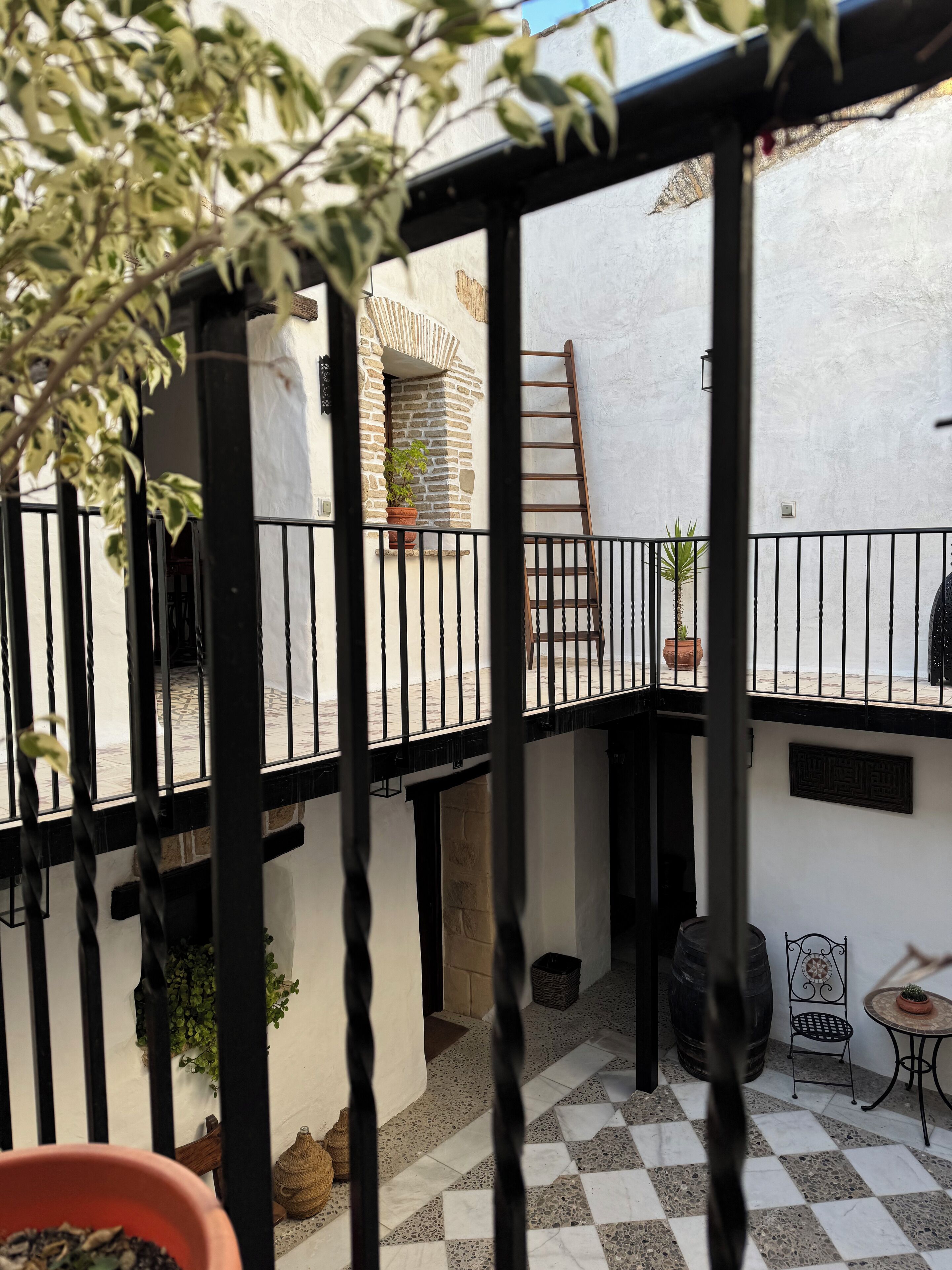 Un patio con suelo a cuadros, una escalera de madera, una planta en maceta y una pequeña mesa con una silla.