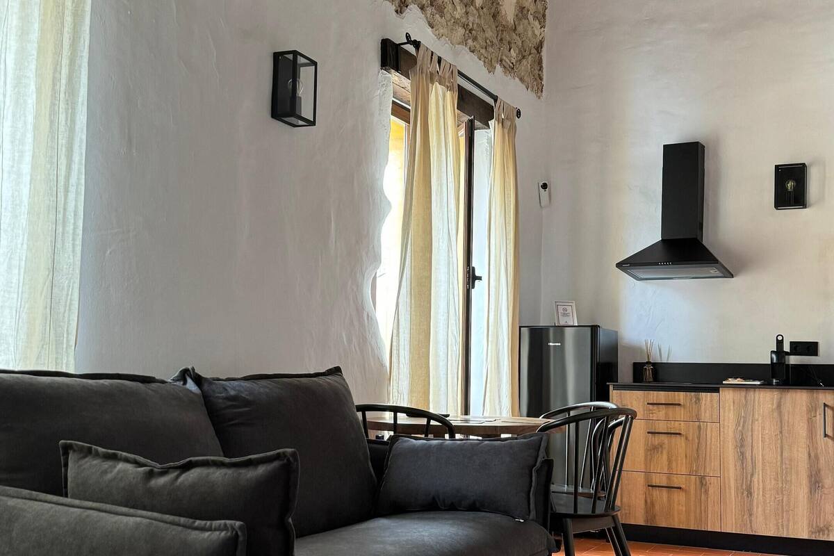 Un salon comprenant un canapé, un fauteuil, un coin cuisine avec une cuisinière et une fenêtre avec des rideaux.