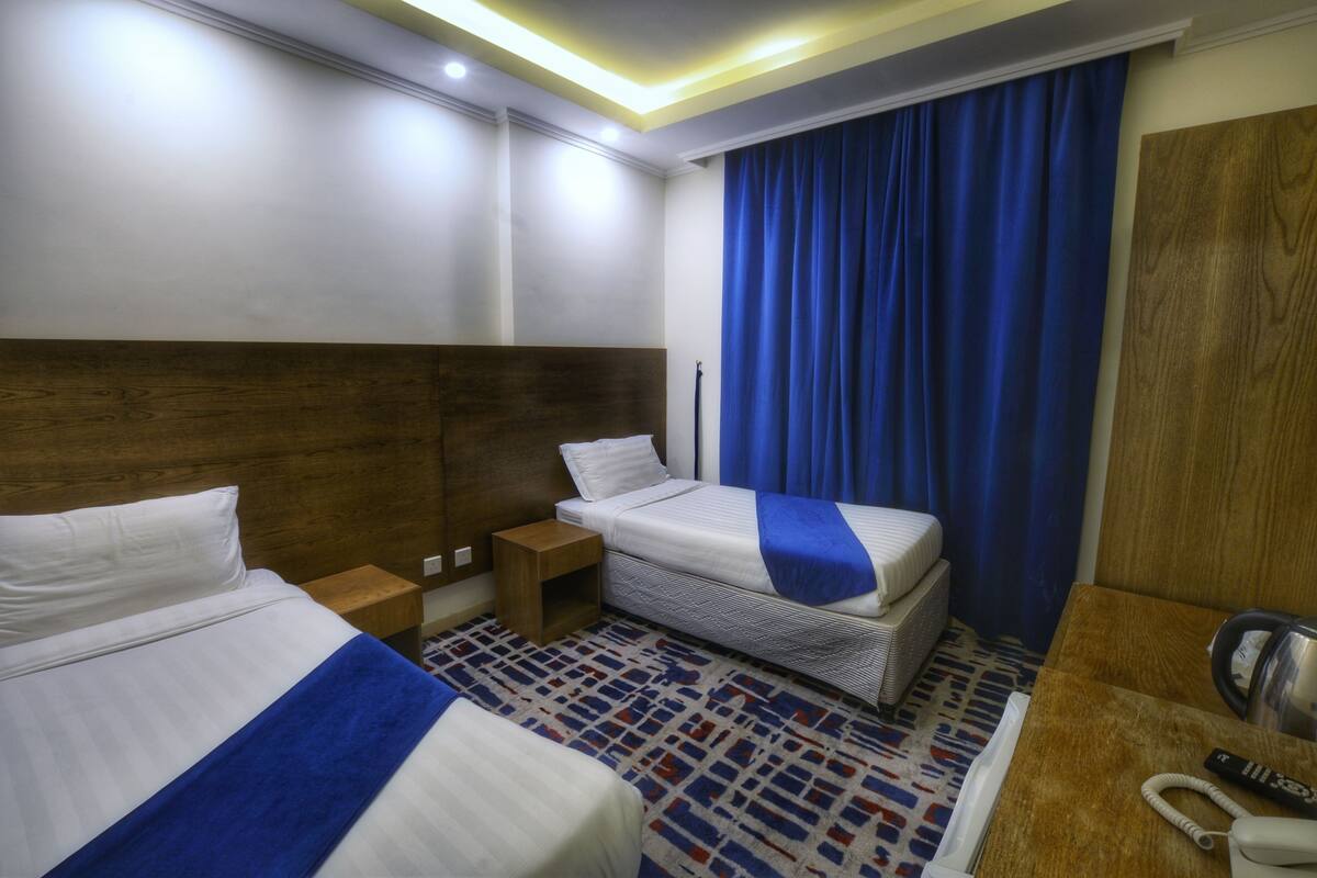 A hotel room with two beds, a wooden headboard, a small wooden nightstand, a patterned rug, a blue curtain, and a wooden desk with a telephone and a kettle.
