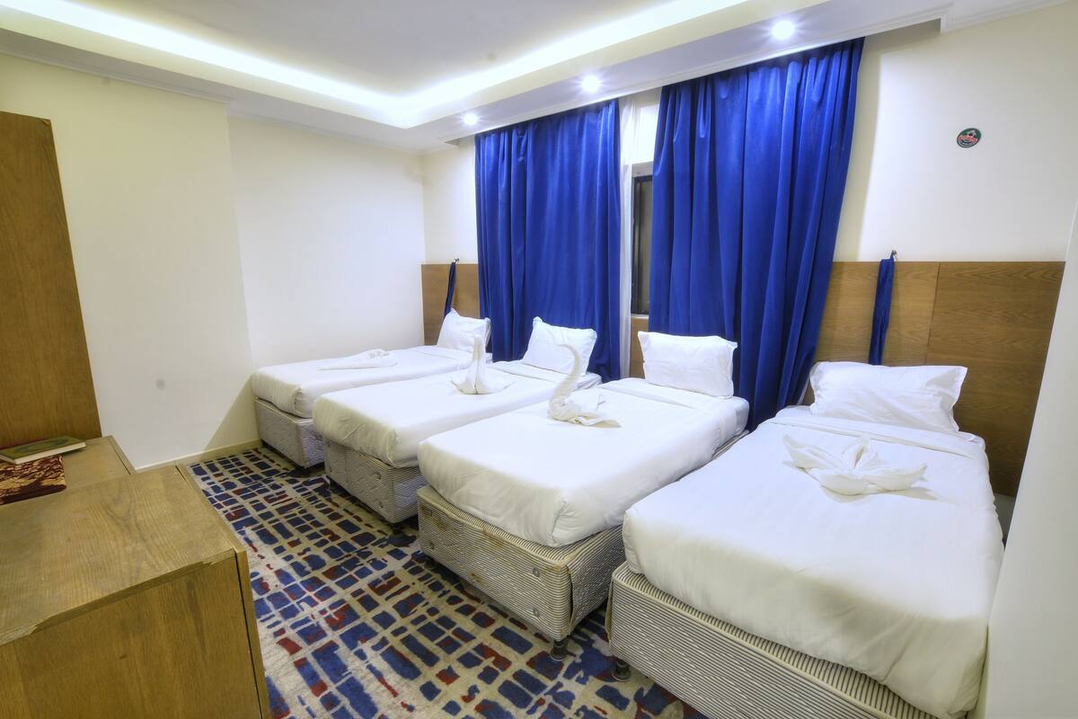 A hotel room with four single beds, a wooden headboard, a patterned carpet, and blue curtains.