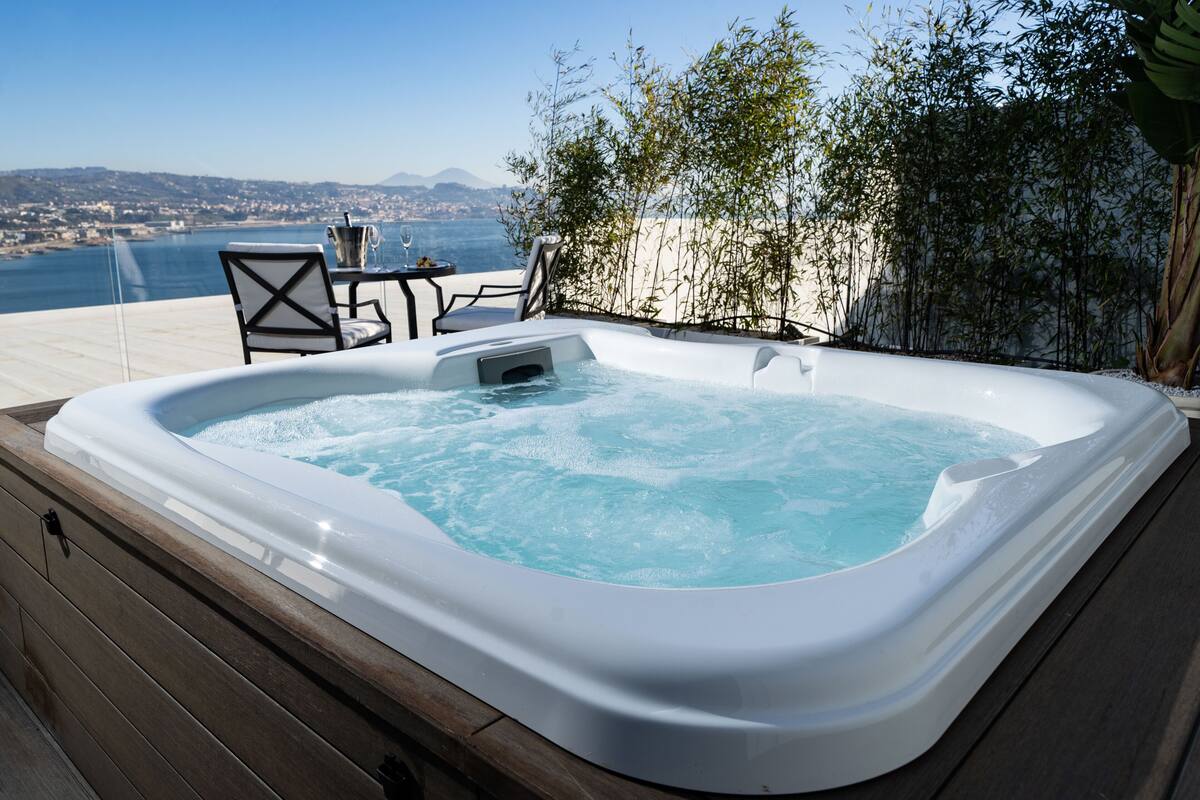 A hot tub with a wooden deck, overlooking a body of water and a distant cityscape.