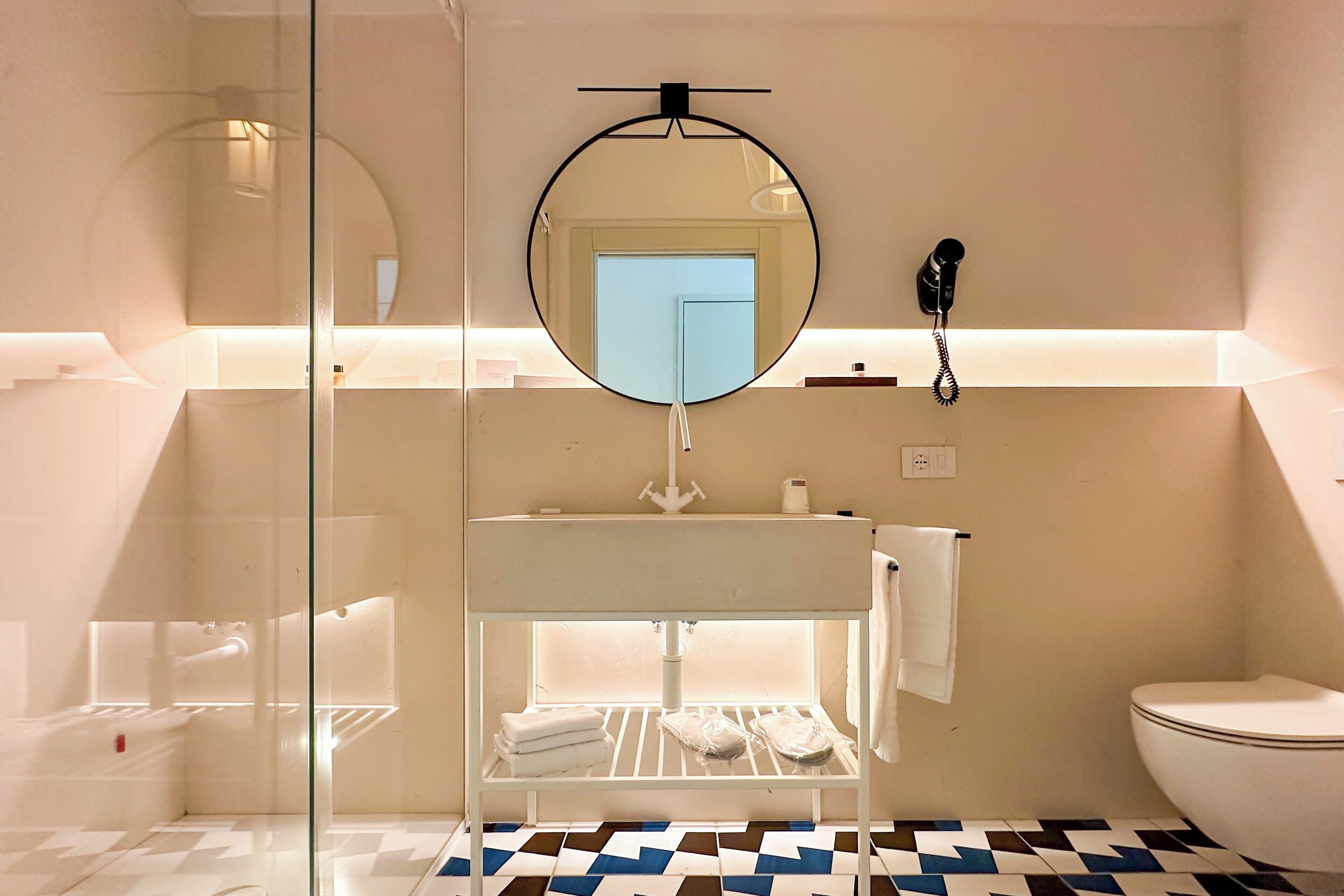Un baño moderno con un lavamanos blanco, un espejo redondo, un teléfono y un inodoro.