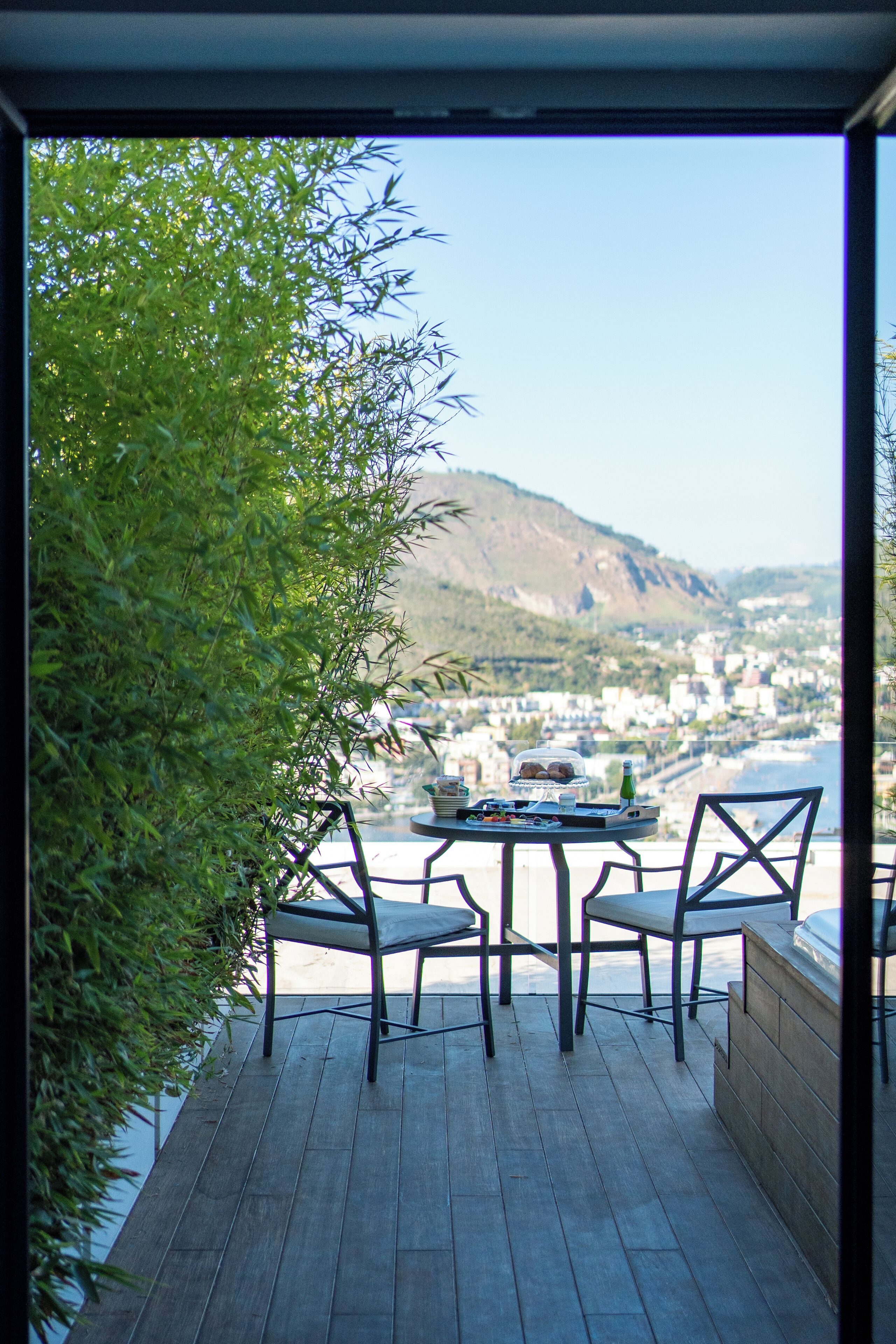 A terrace with a table and chairs, overlooking a coastal town and hills.