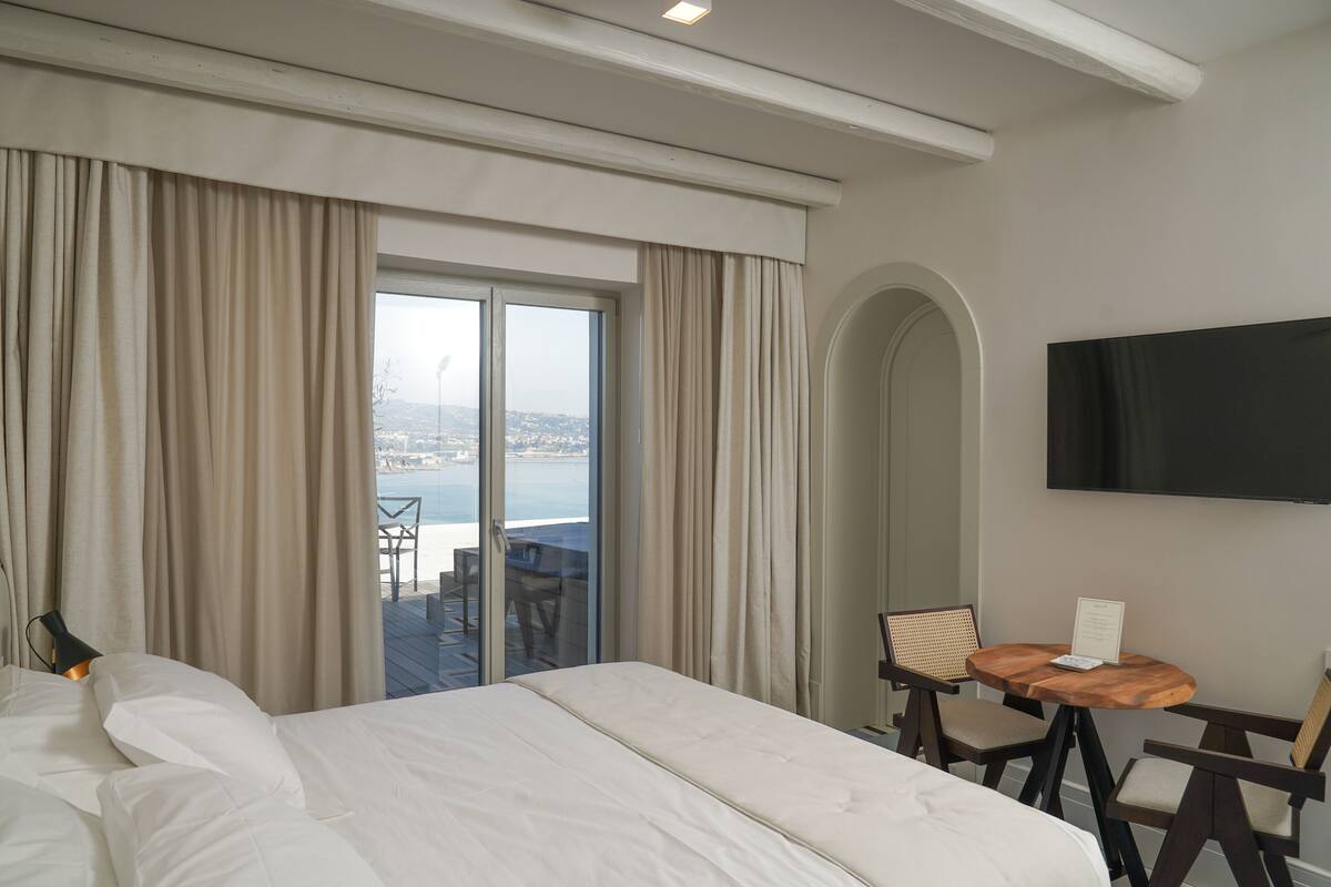 A hotel room with a large bed, a TV mounted on the wall, a small table with a chair, and a balcony with a view of the sea.