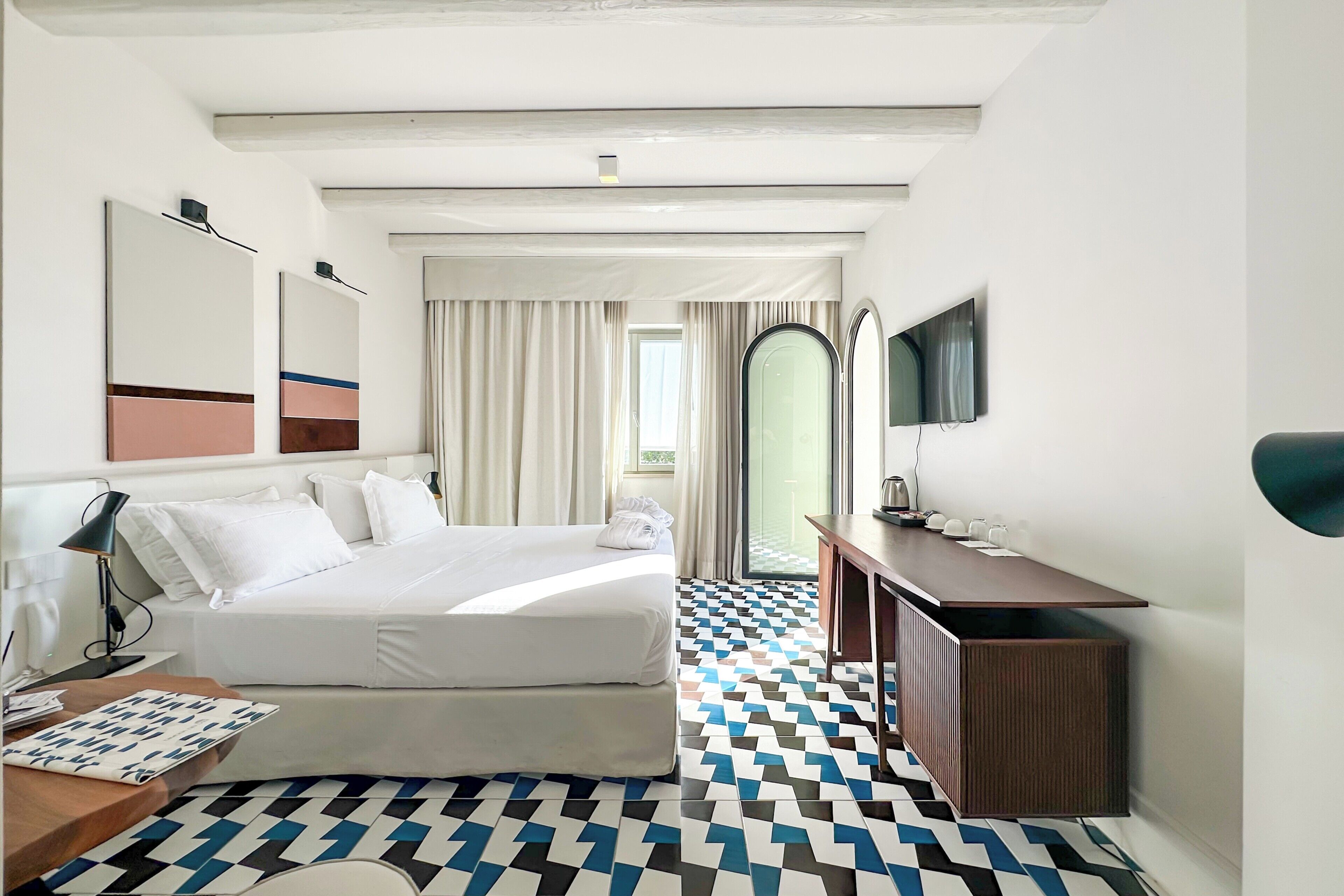 A hotel room with a large bed, a desk with a computer, a TV, and a patterned floor.