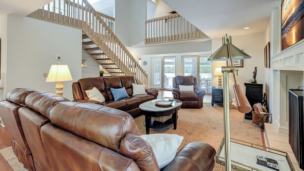 A spacious living room with a leather sofa, a coffee table, and a fireplace. A wooden staircase leads to an upper level. - 2 Duck Hawk - Sea Pines just a Short Walk to the Beach & Private Pool (Hilton Head Island)