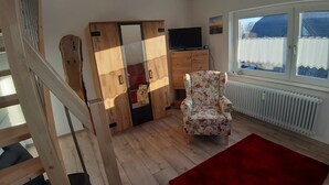 A room with a wooden wardrobe, a floral armchair, a red rug, a television, and a window with curtains. - Ferienwohnung "weit Blick" (Dornum)