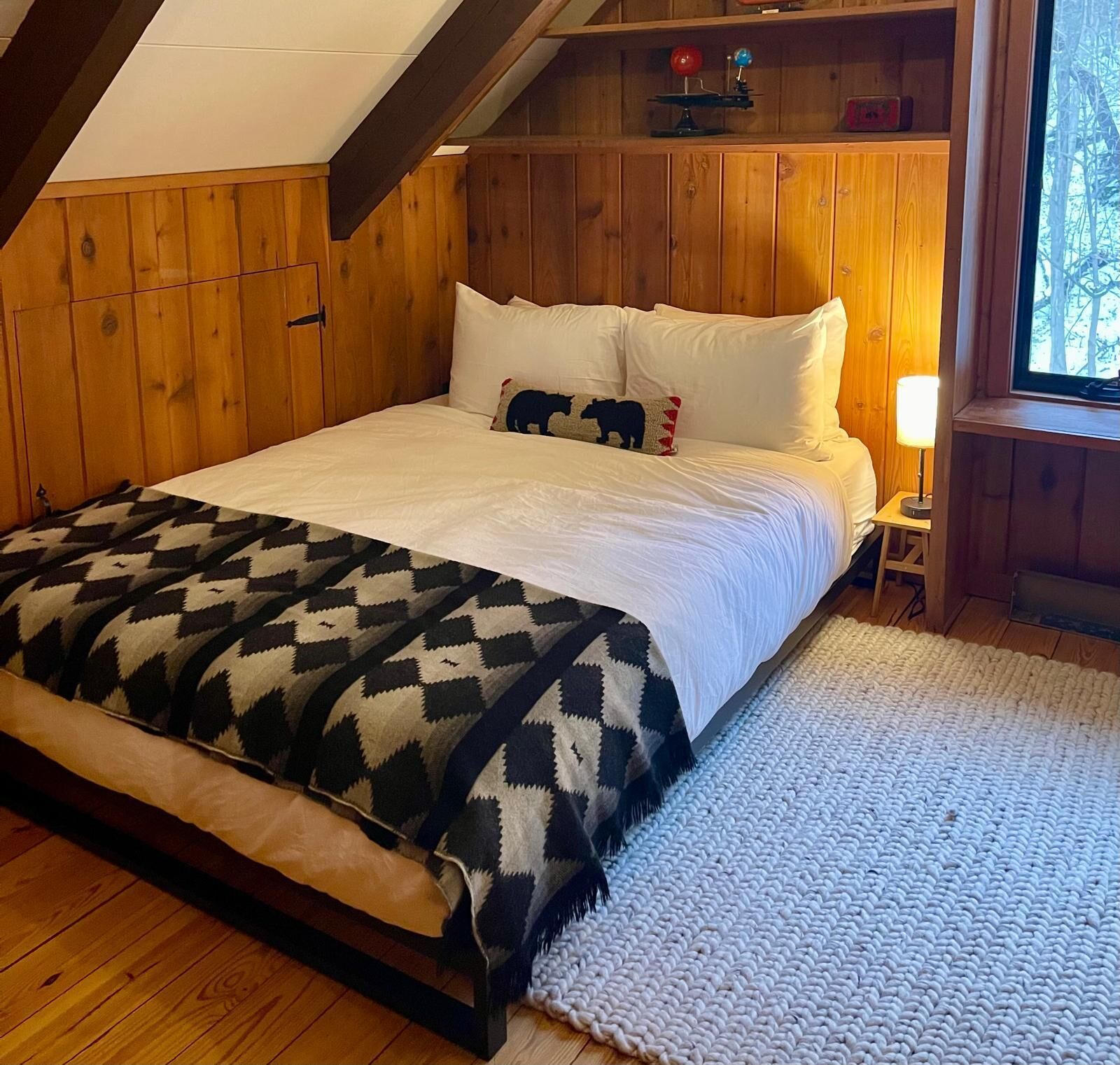 Une chambre avec un plafond en bois, un lit recouvert de draps blancs, un plaid à motifs et une table de chevet avec une lampe.