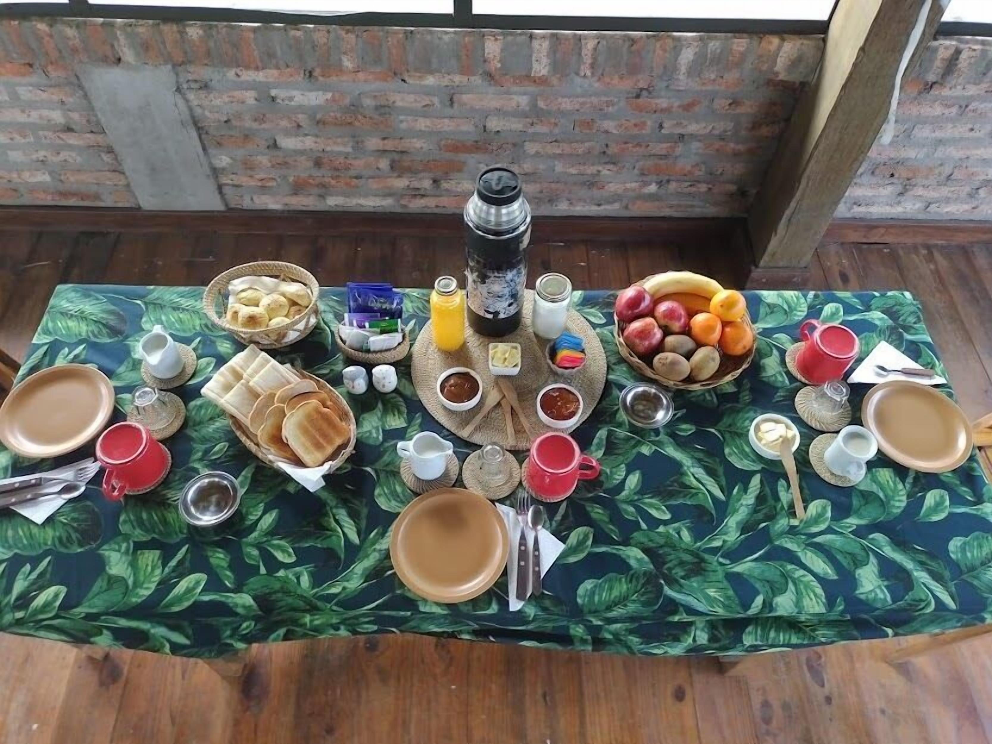 Desayuno con pan, frutas y bebidas sobre una mesa con mantel estampado de hojas tropicales.