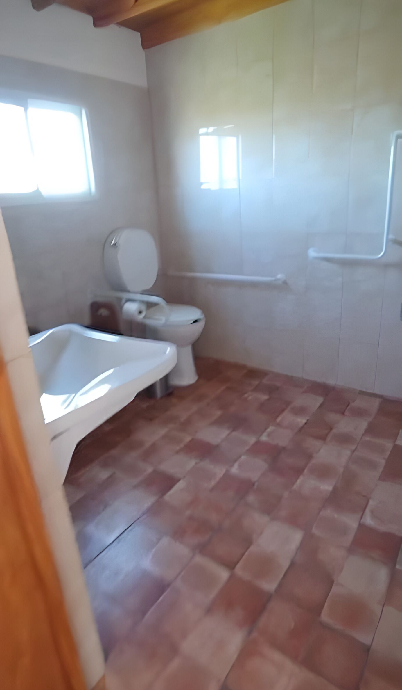 A bathroom with a white toilet, a white sink, and a shower. The floor is tiled, and there are windows allowing natural light.