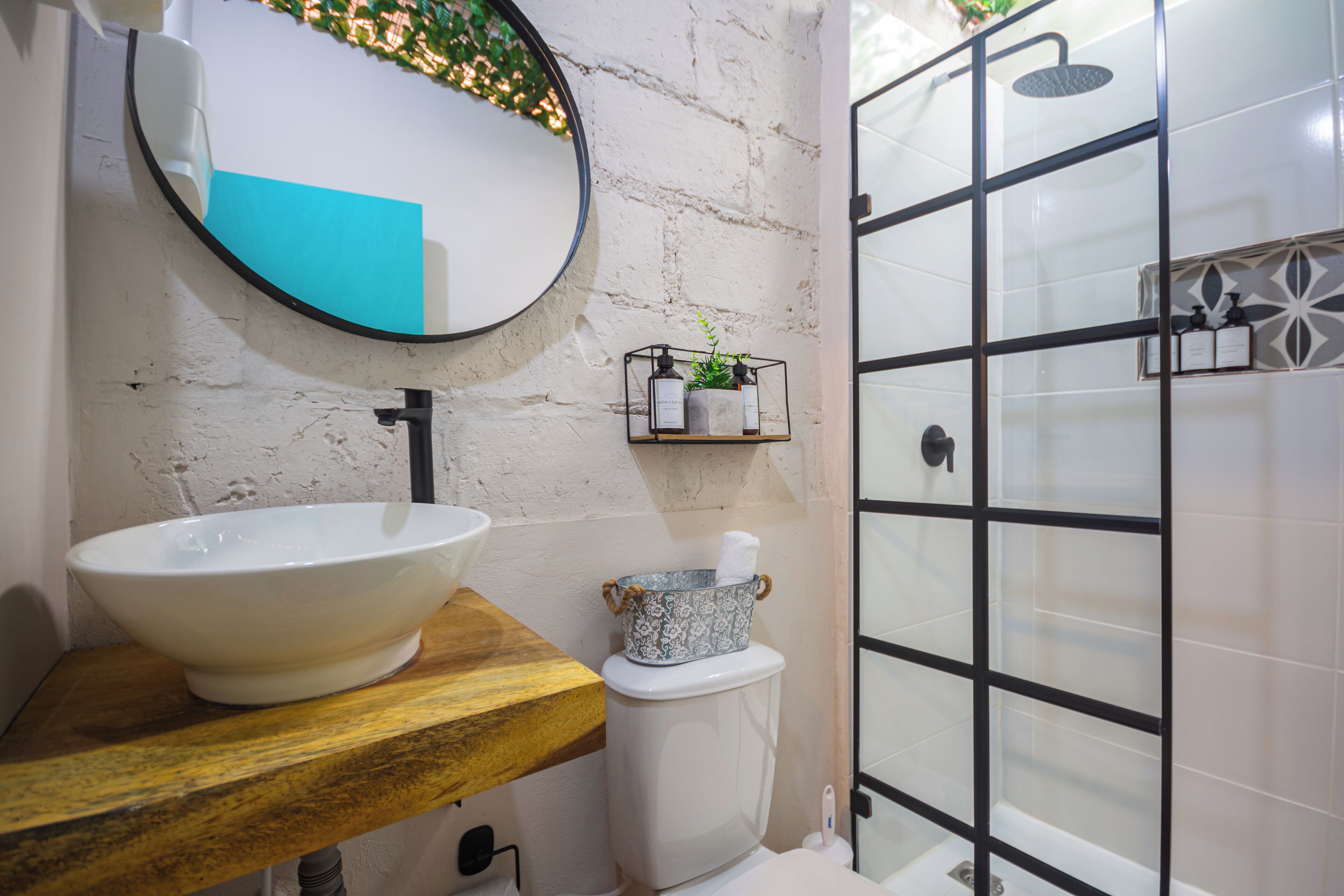Une salle de bain avec un lavabo blanc, un plan de toilette en bois et un miroir rond. On y trouve une petite étagère avec des articles de toilette et une plante en pot.