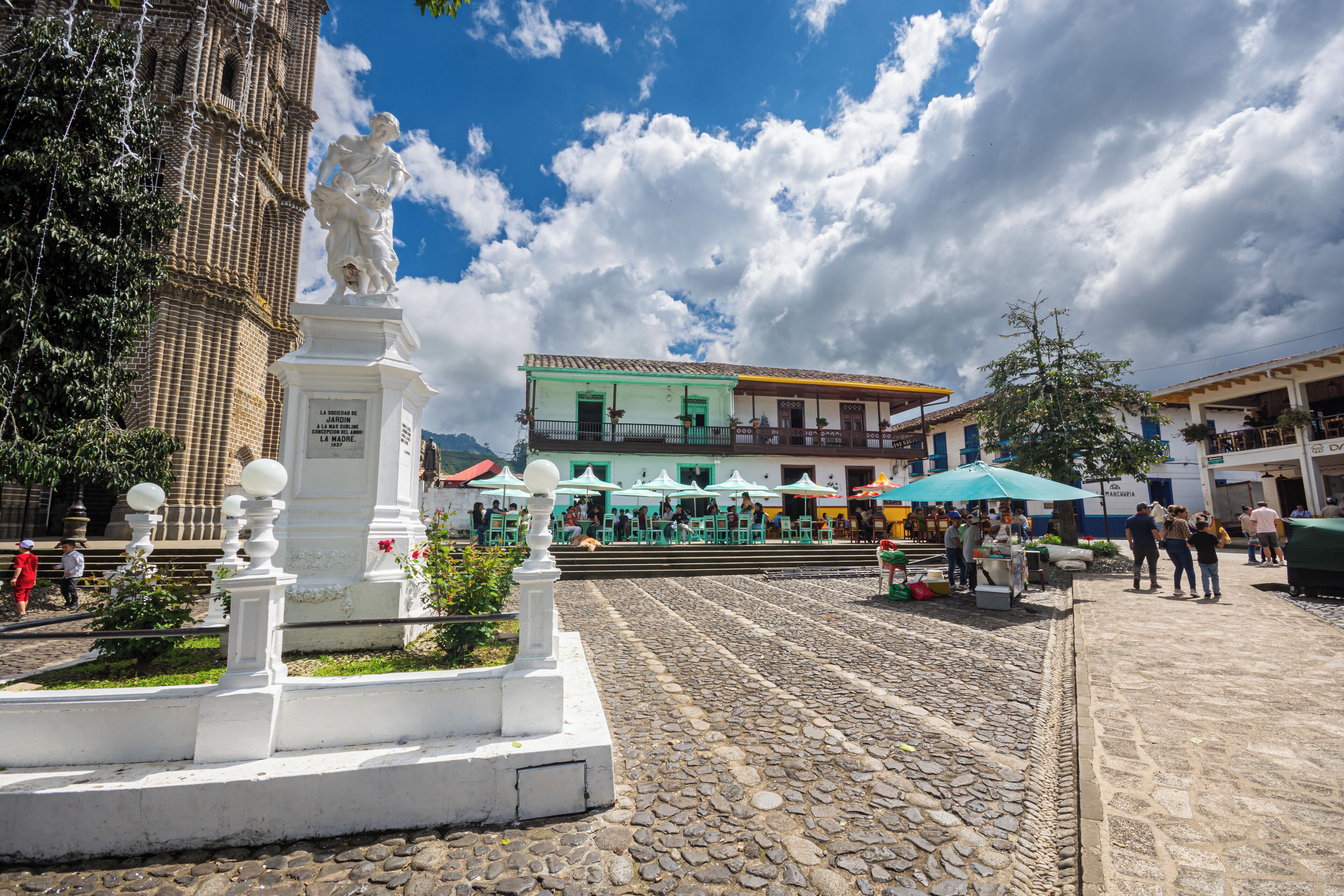 Uma praça de paralelepípedos com uma estátua, uma torre de igreja e edifícios com mesas ao ar livre.