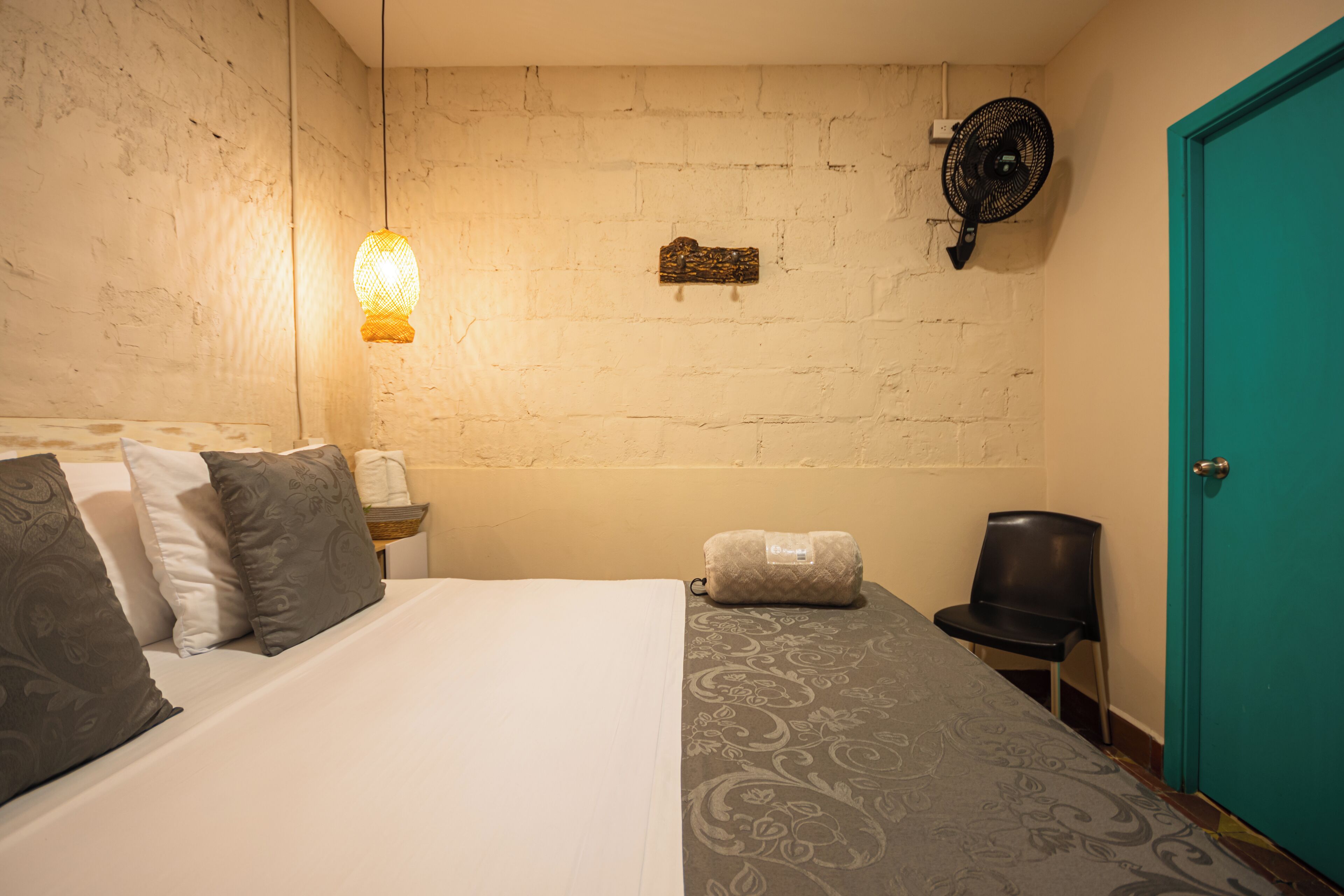 Un dormitorio con cama, almohadas, una mesita de noche, un ventilador de pared y una puerta color turquesa.