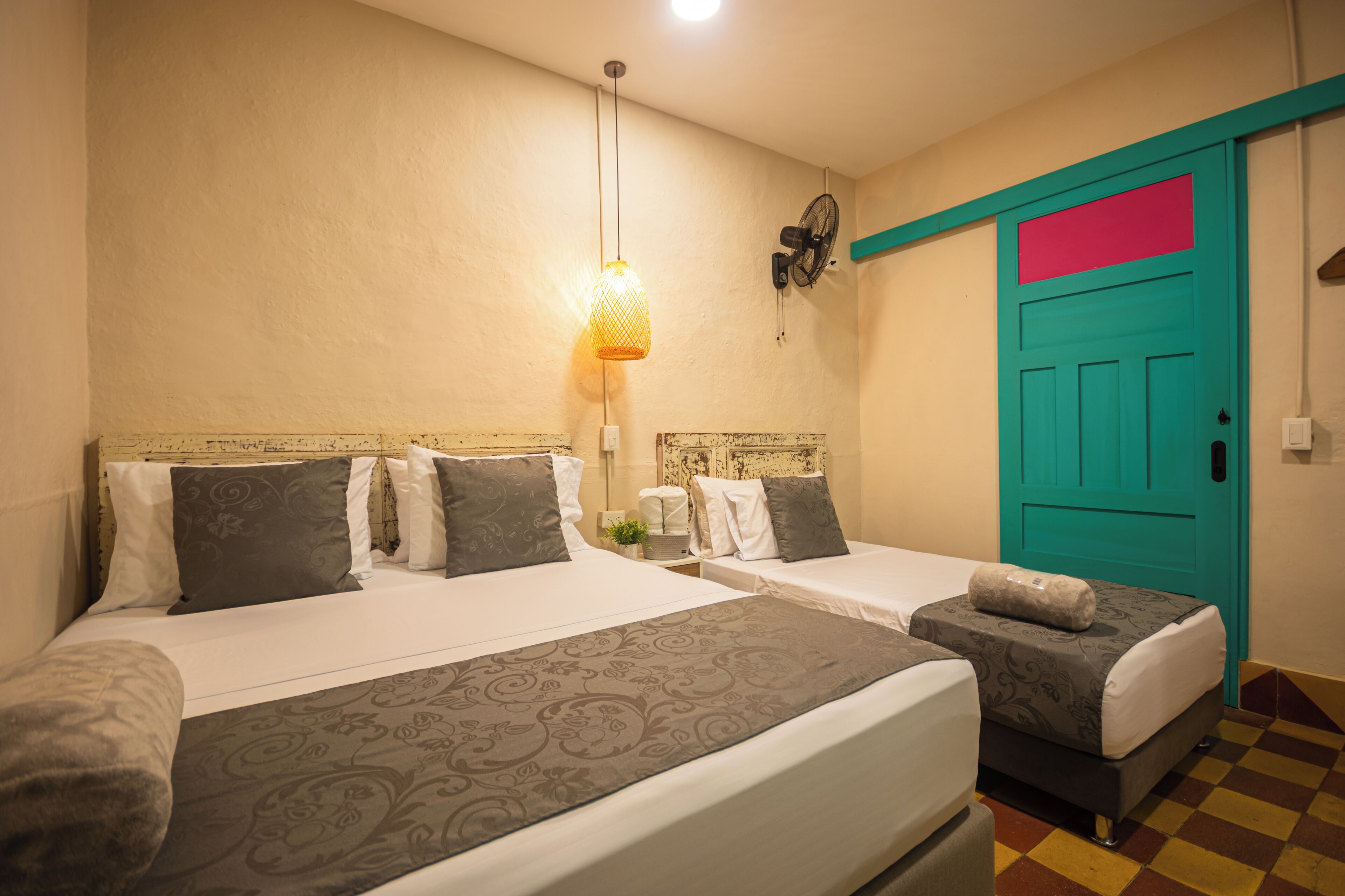 A hotel room with two beds, a teal door, a wall-mounted fan, and a hanging light fixture.