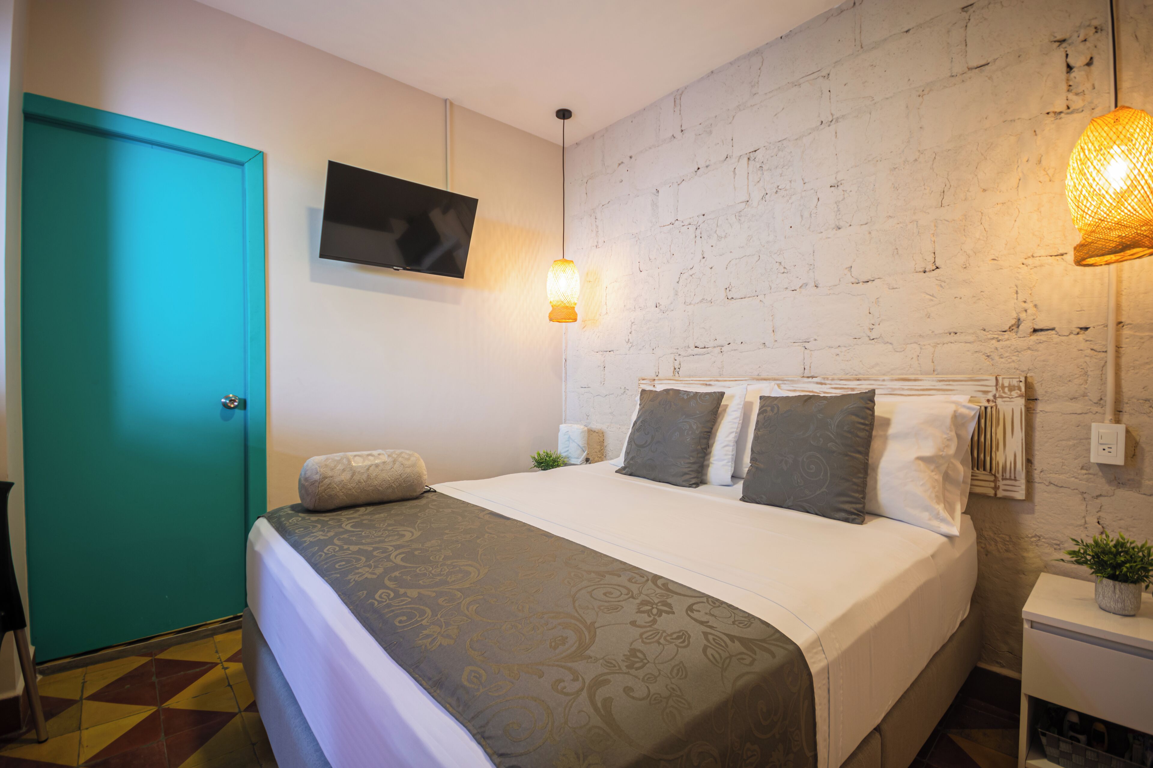 A hotel room with a bed, a television mounted on the wall, a teal door, and a brick wall.