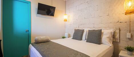 A hotel room with a bed, a television mounted on the wall, a teal door, and a brick wall.