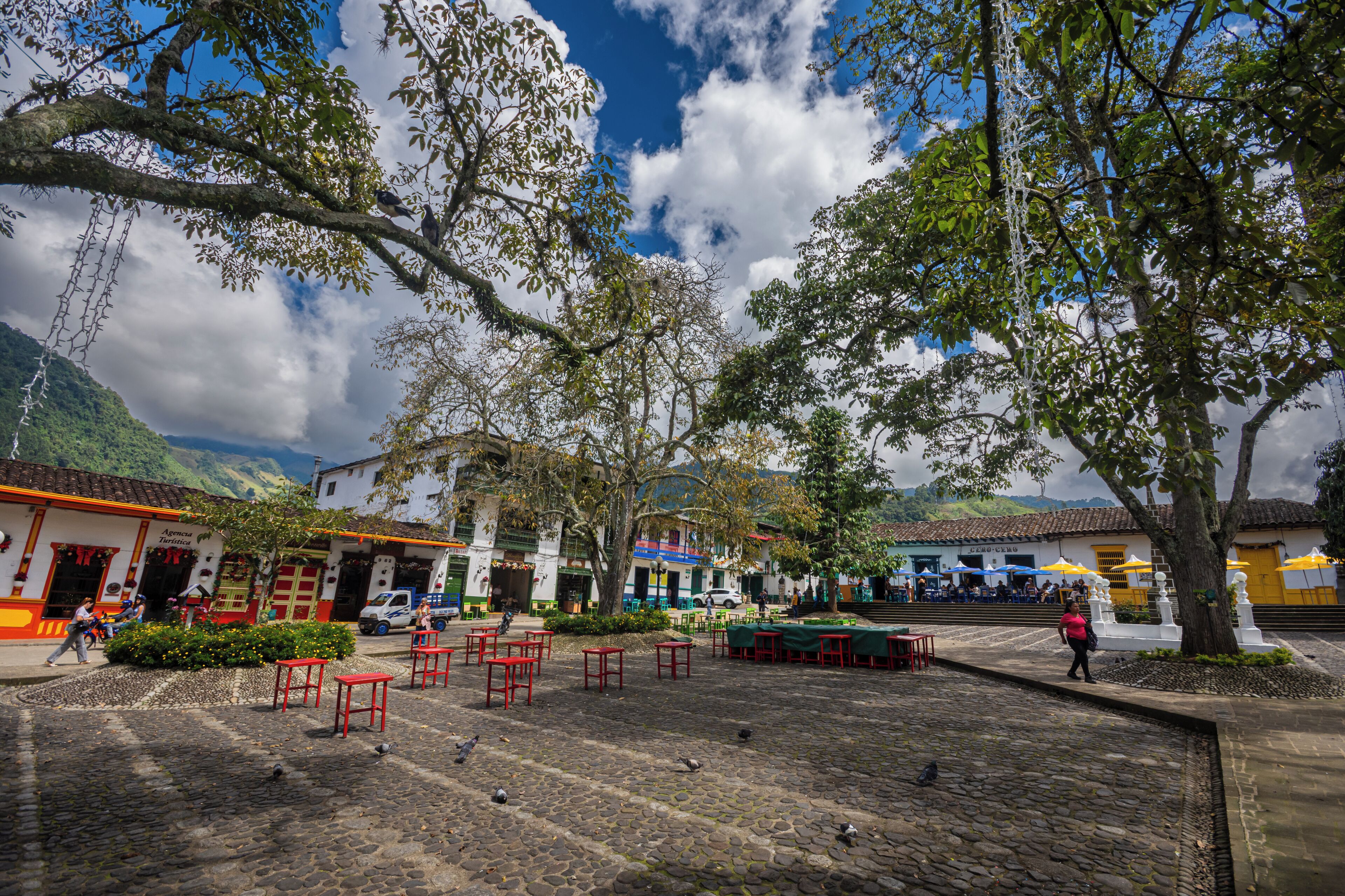 A cobblestone plaza with outdoor seating, trees, and buildings with colorful facades.